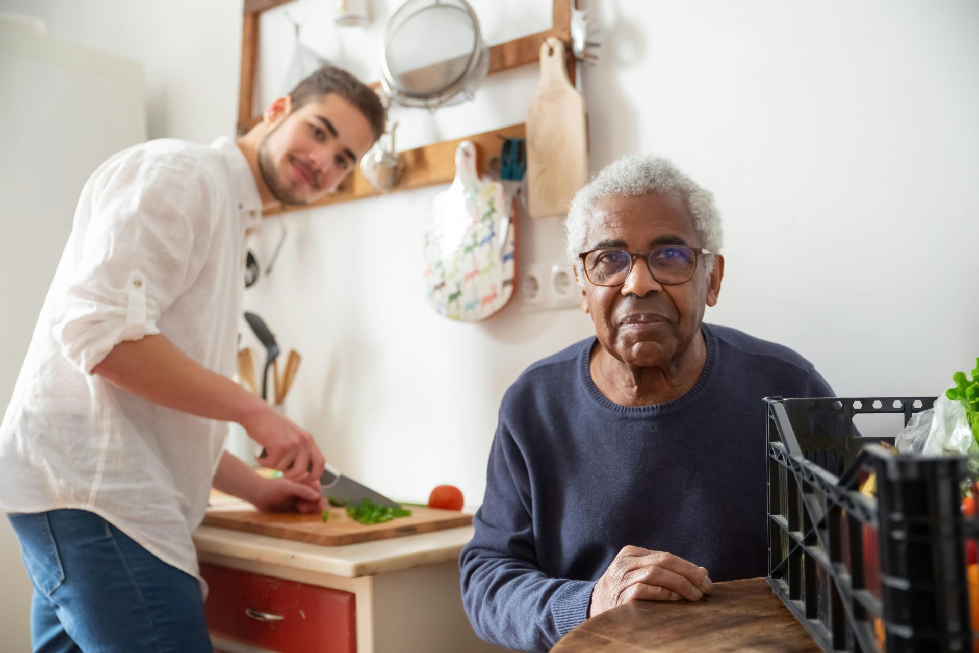 Un jeune adulte aide une personne âgée dans la cuisine, illustrant l'importance des soins et du soutien à domicile.